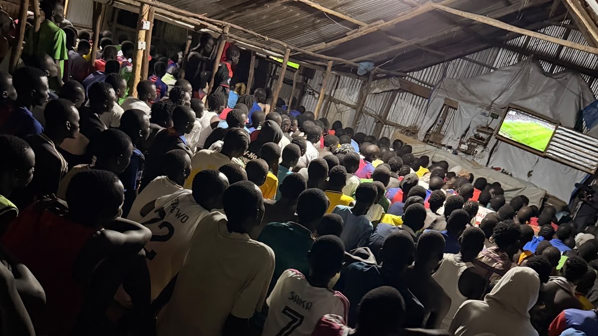 A packed video hall under a tin roof, everyone facing a screen showing a football match